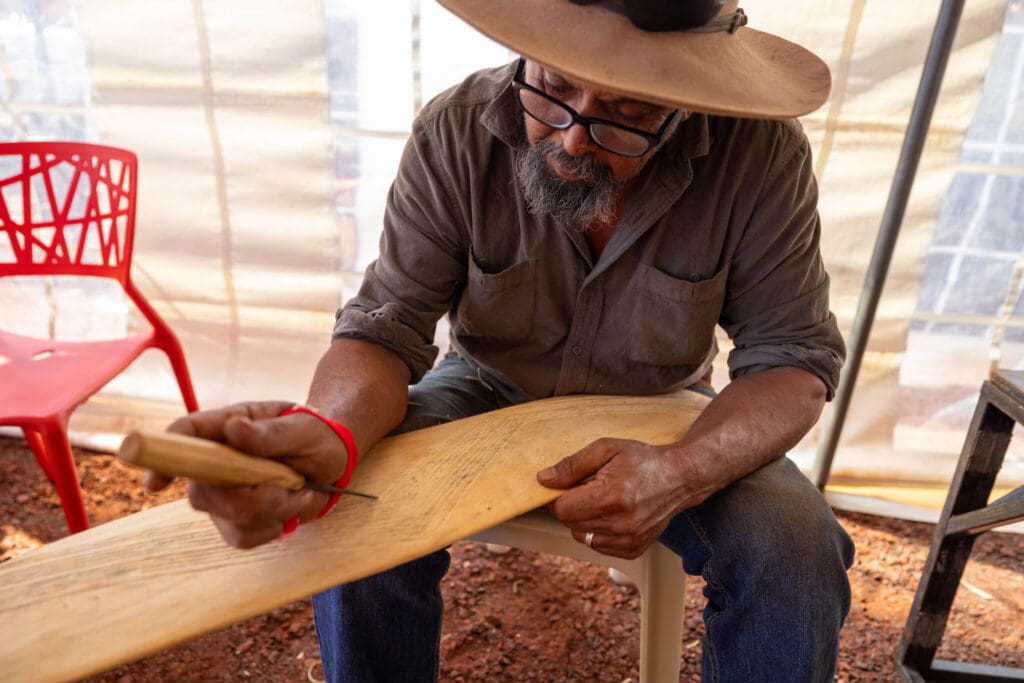 Banjima cultural heritage practitioner carving a wooden object by hand in a shaded work area